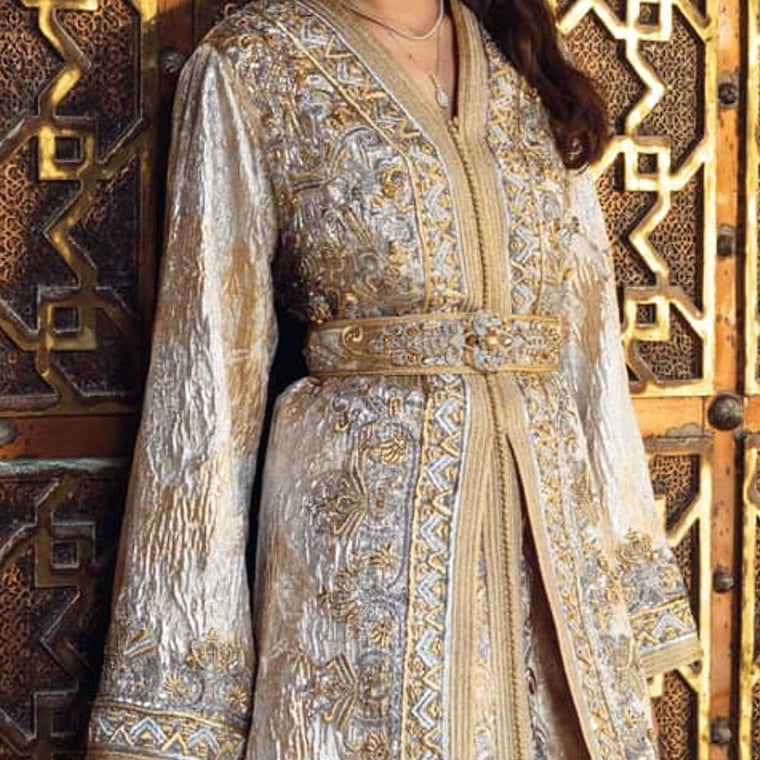 Pearl Gate ivory and silver embroidered Moroccan kaftan with a beaded belt, before an engraved brass door
