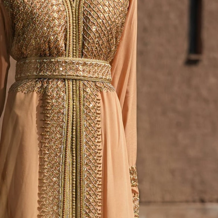 Apricot crepe Moroccan kaftan with a gold sequin plastron and embroidered belt, against a Marrakech adobe wall